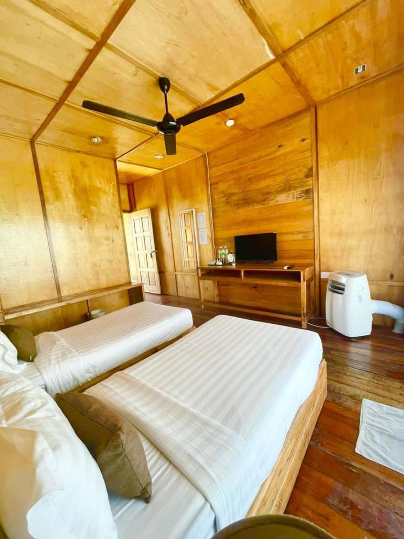 A charming bedroom at Nusakuya Resort near Bum Bum Island, featuring two single beds, a modern TV desk, warm wooden plank walls, varnished timber flooring, and refreshing AC.