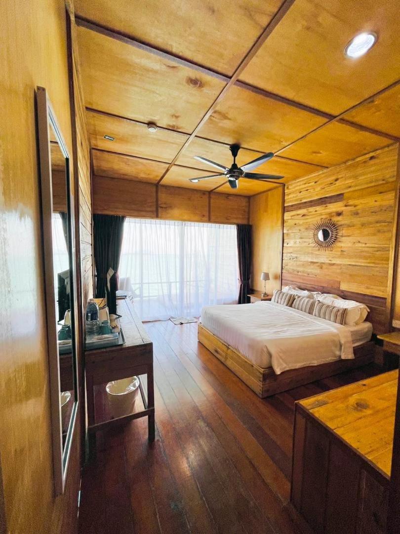 A relaxing bedroom at Nusakuya Resort featuring a bed facing seamless glass doors opening onto the balcony, rich wood paneling with polished timber floors, and AC.