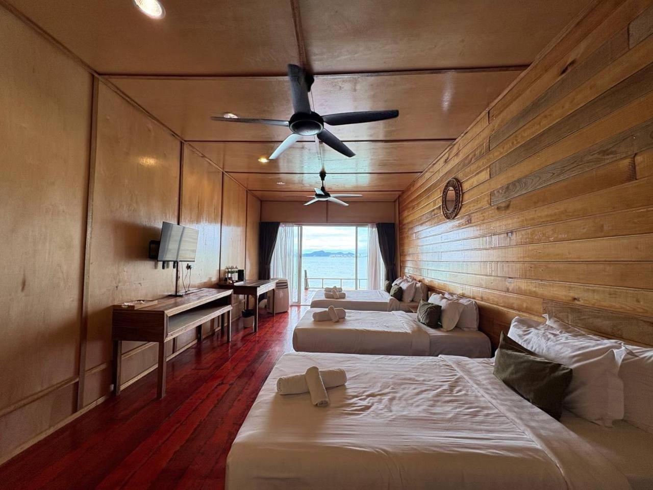 A spacious bedroom at Nusakuya Resort featuring three beds facing seamless glass doors to the balcony, rustic timber walls and AC.
